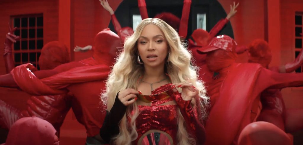 Beyonce rocking blonde hair dressed in a crimson red body suit surrounded by a group of dancers also dressed in crimson red.
