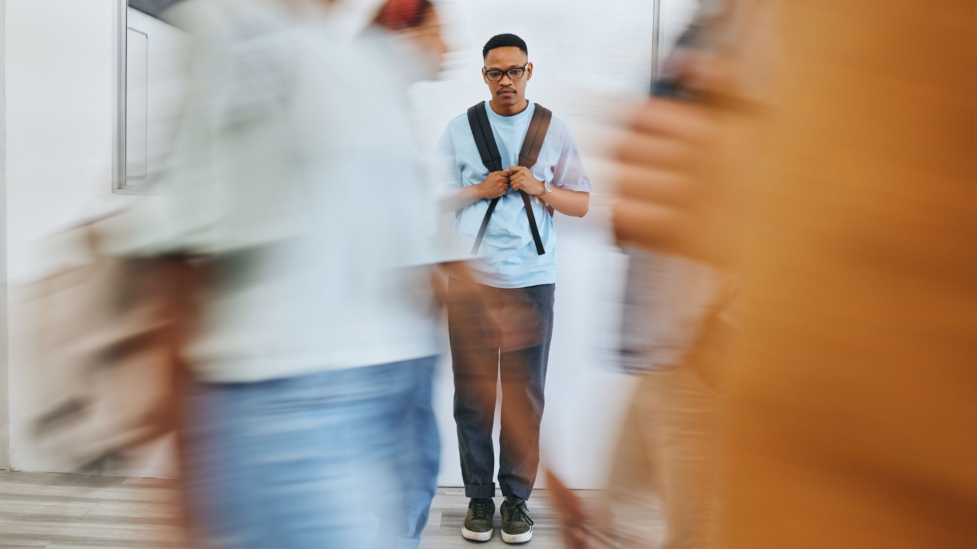 Young Black man with mental health challenges is walking down a hall in a blue shirt and jeans carrying a backpack while the people around him are blurred.