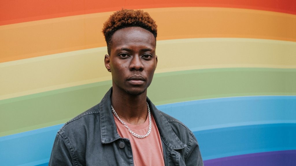 Black male member of the LGBTQIA+ community standing in front of a rainbow wall.