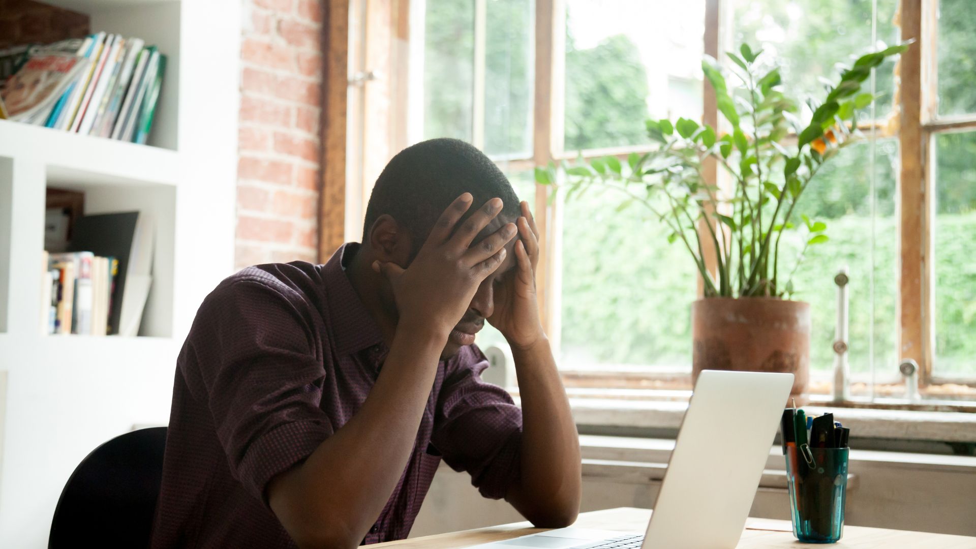 Frustrated Black business man in purple button-up t-shirt holding his head in anguish in front of his computer screen in his home office due to brand safety bias.