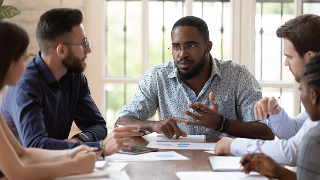 A group of diverse business men and women dressed in business casual attire sitting around a board room table in deep discussion about brand safety bias. 