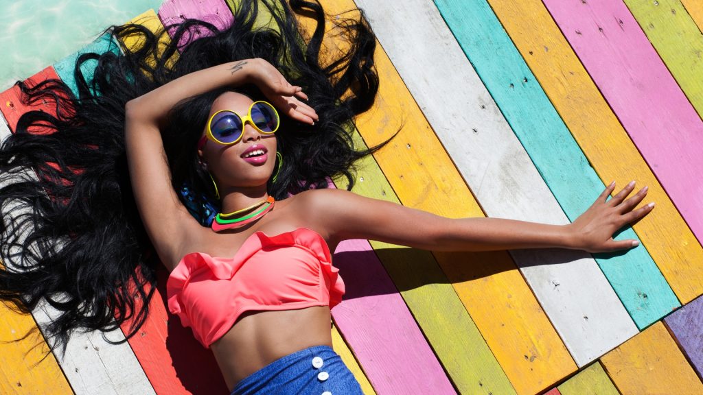 Young Black woman laying out on a brightly colored beach towel in a bright pink bikini top and royal blue short shorts - in promotion of Travel Noire City Guides