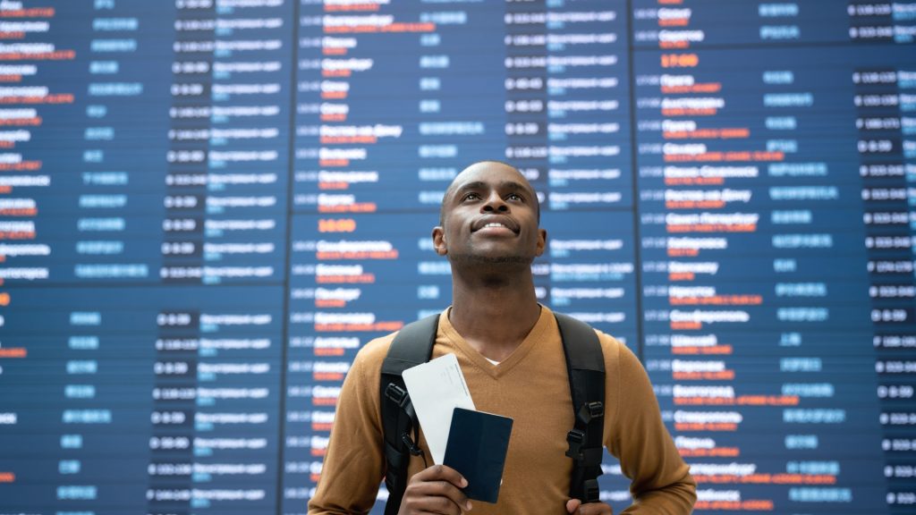 Black male traveler at the airport standing in front of an information board showcasing incoming and outgoing flights - in promotion of Travel Noire City Guides