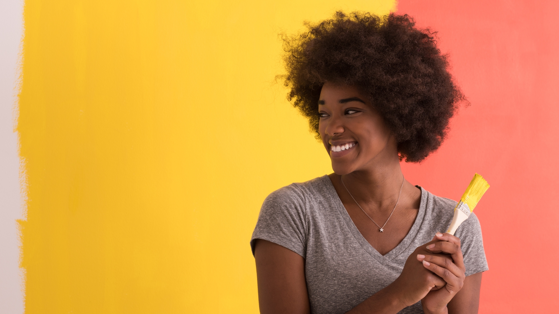 African American woman with an afro holding a paint brush in front of a two-toned wall that she just painted.