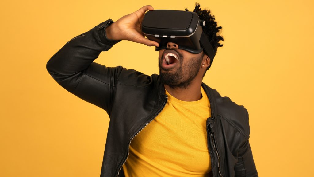 Black male gamer wearing a VR headset in awe of in-game advertising and standing against a yellow backdrop 