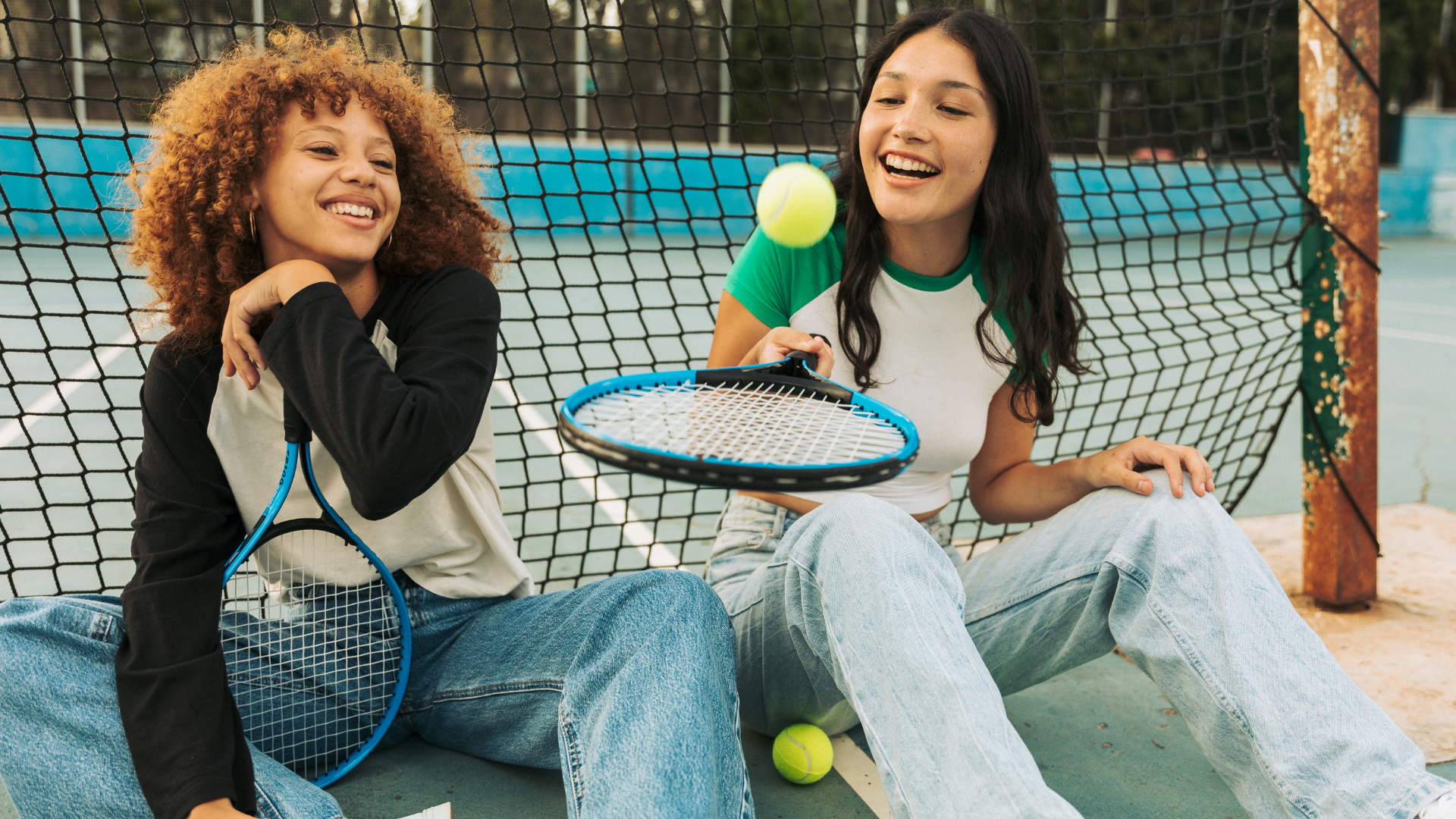 2 multicultural teen girls sit together on a tennis court laughing with one another.