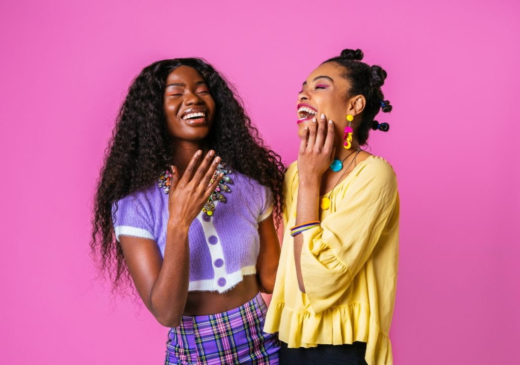 Two happy and smiling women of color wearing vibrant colors standing in front of a bright pink background celebrating Women's History Month.
