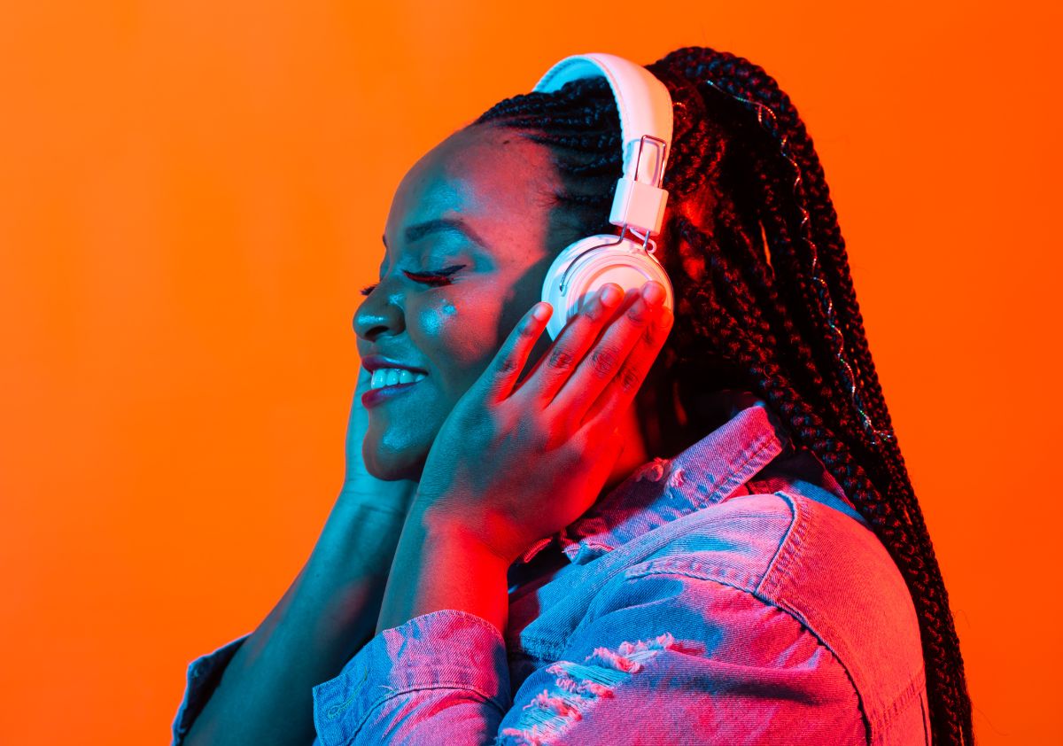 A smiling Black woman with box braids wearing headphones and smiling in front of an orange backdrop as she prepares for the Blavity House Party Music Festival.