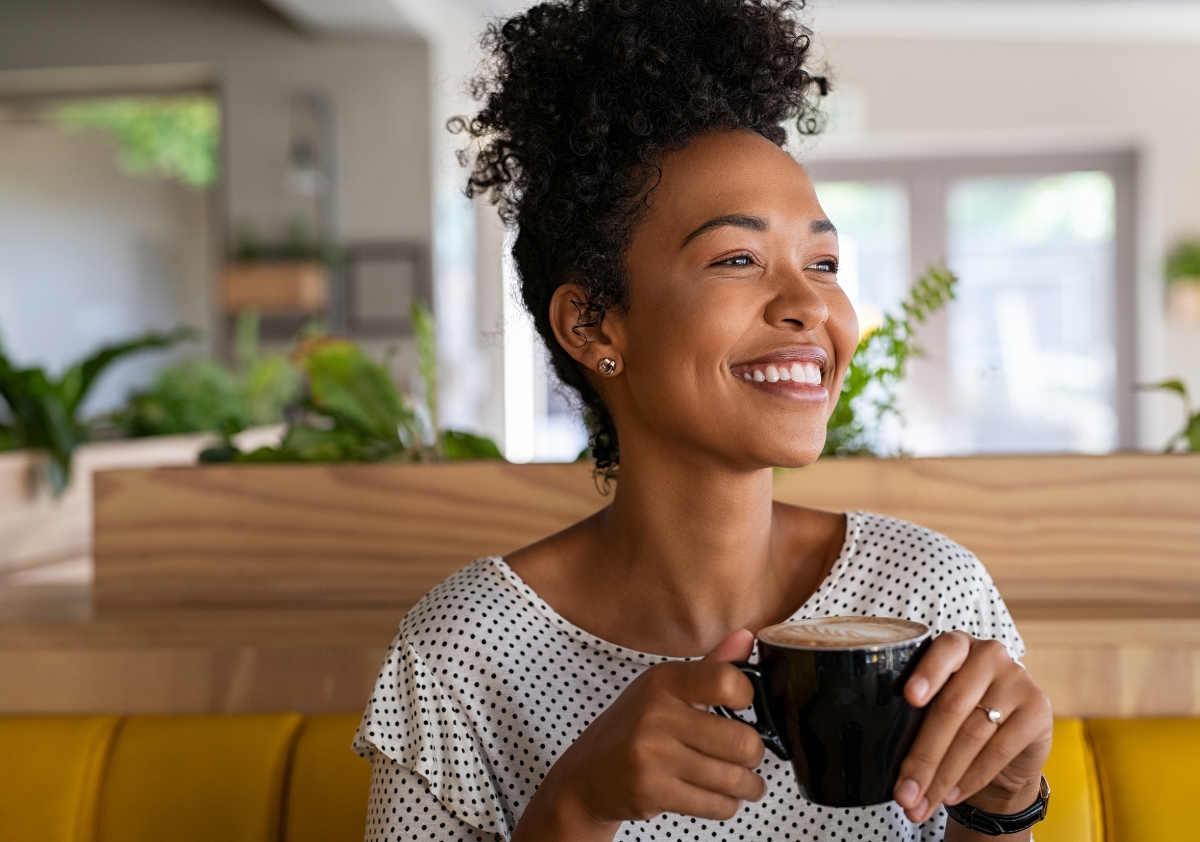 Joyful Black woman with curly hair pulled up into a high bun smiling while holding a fresh cup of coffee