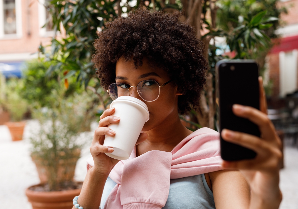 A young black woman with curly hair drinking a cup of coffee while taking a selfie for the Creator Collective