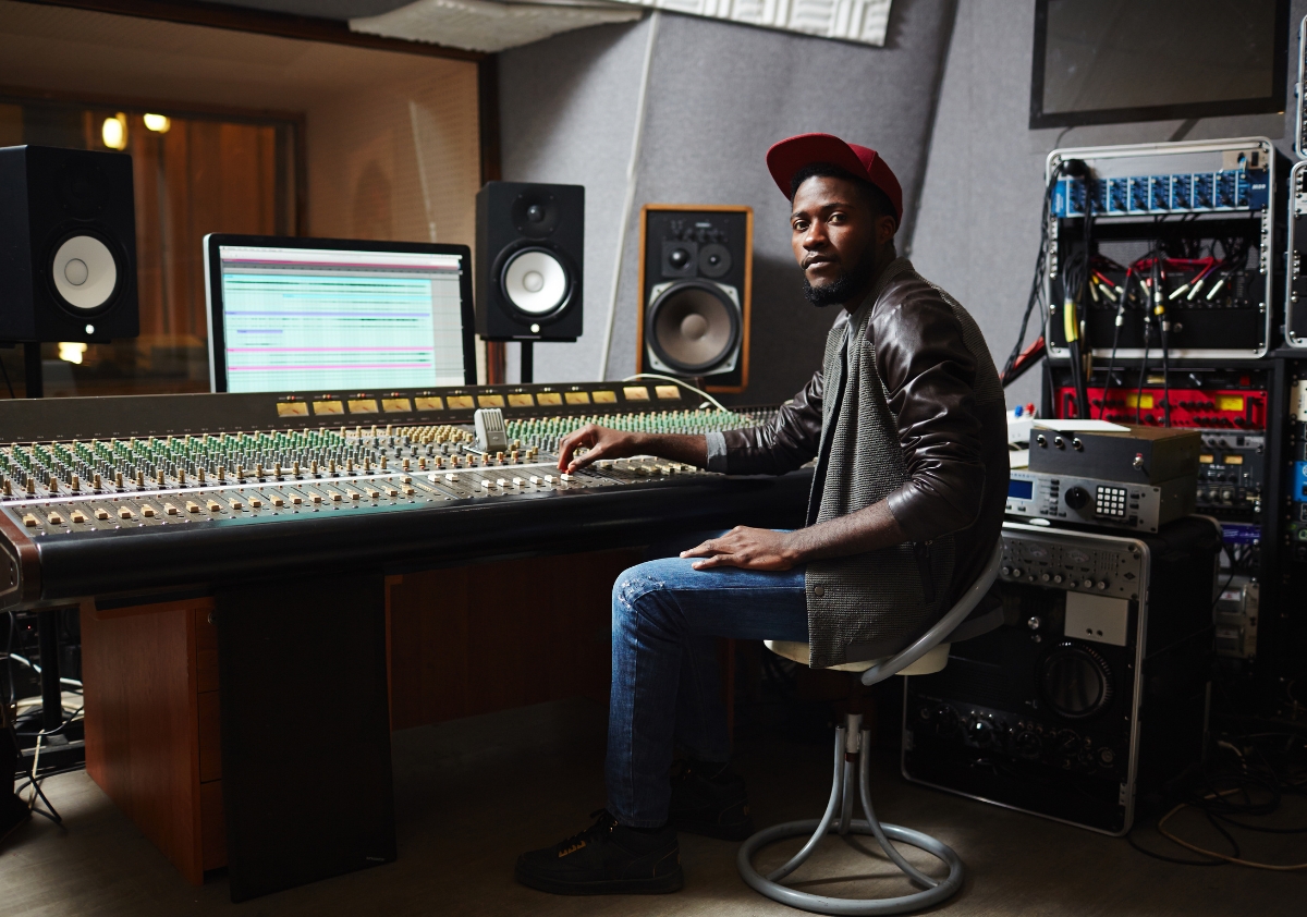 Black male audio engineer/producer wearing a baseball hat, a jacket, and blue jeans sits in the studio thinking about how to support diverse talent