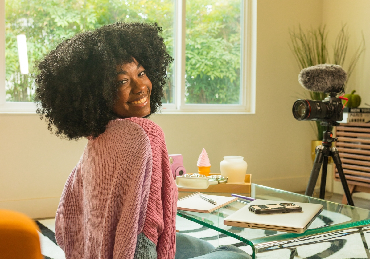 A female influencer of color with a beautiful afro wearing a pink sweater sits in front of a camera creating brand storytelling content for a client