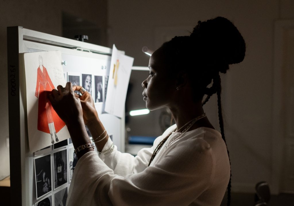 A black female designer with braids wearing a white t-shirt pins up a sketch of her latest apparel concept late night in the studio while also contemplating the need to support diverse talent