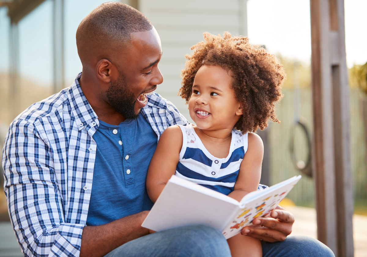 Black father featured in inclusive marketing campaign sitting on the porch with his toddler daughter reading a book.