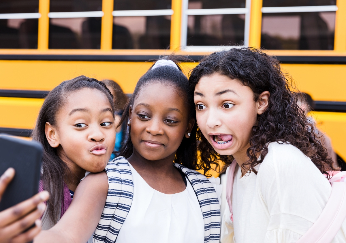 3 vibrant Black tween girls take a selfie in front of a yellow school bus