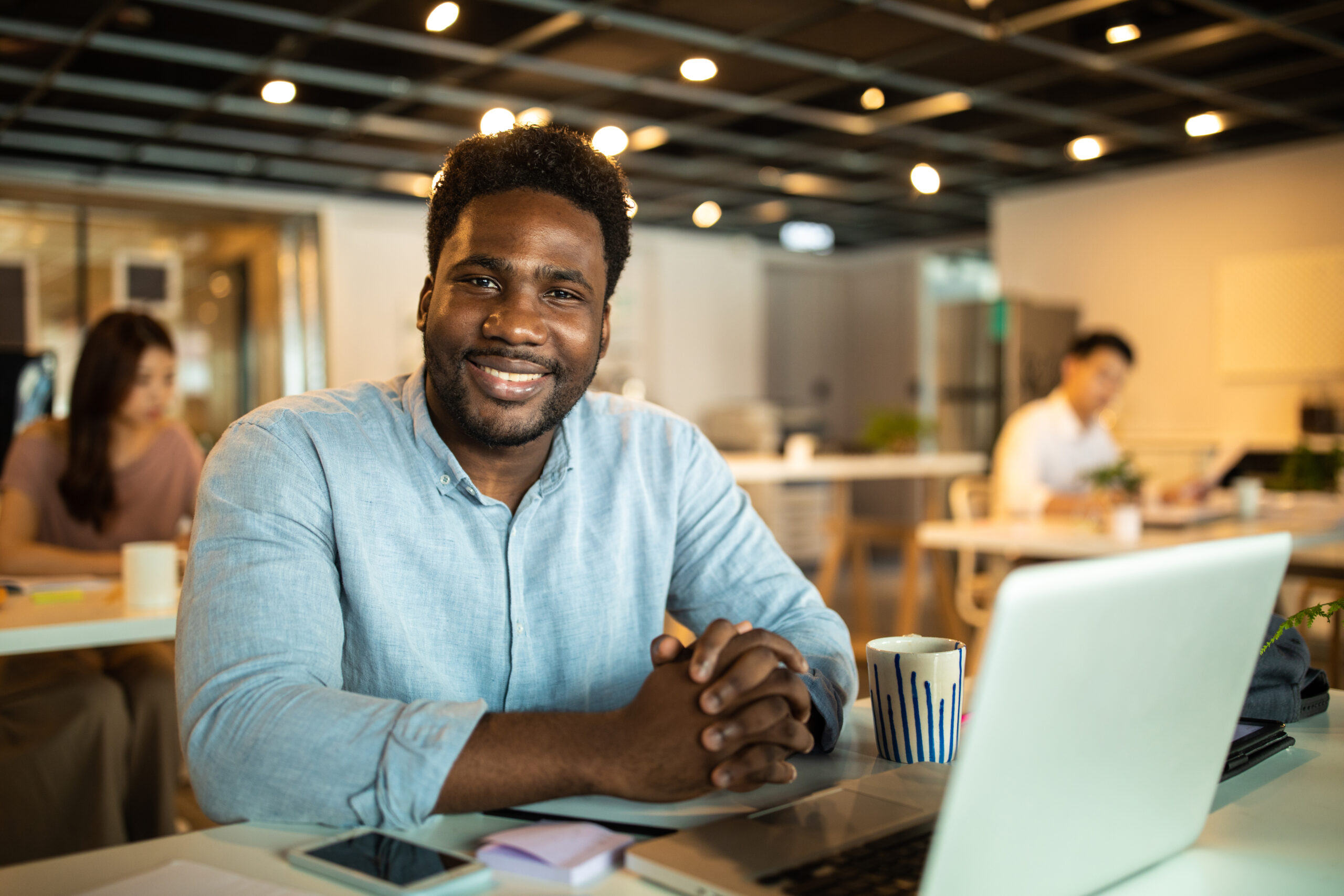 A young man with a short beard and curly hair is sitting at a desk, smiling and looking at the camera. He is wearing a light blue button-up shirt. On the desk, there is a laptop, a smartphone, a coffee mug, and some documents. The background shows a modern, open office space with colleagues working at desks, under soft, warm lighting. The atmosphere is professional and welcoming.