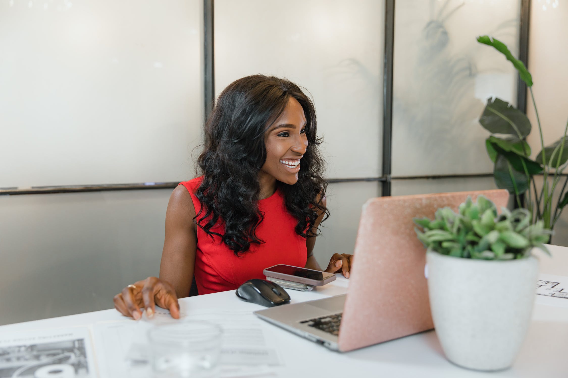 A young woman with long, curly hair is sitting at a desk, smiling and looking at her laptop. She is wearing a sleeveless red top. On the desk, there is a smartphone, a computer mouse, some documents, and a potted plant. The background features a glass partition with plants, creating a bright and modern office environment.