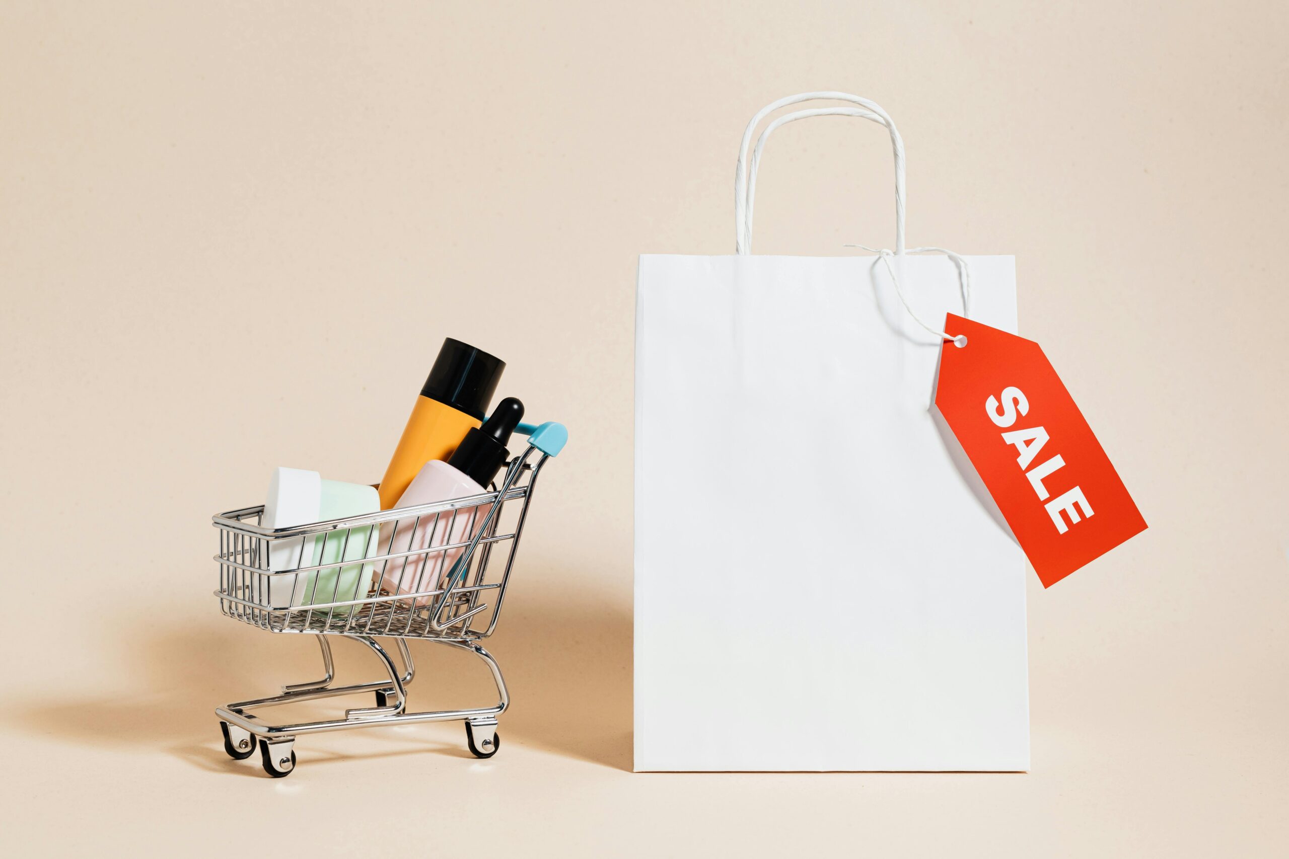 Miniature shopping cart filled with cosmetic products next to a white shopping bag with a red 'SALE' tag, symbolizing product marketing and promotional sales.